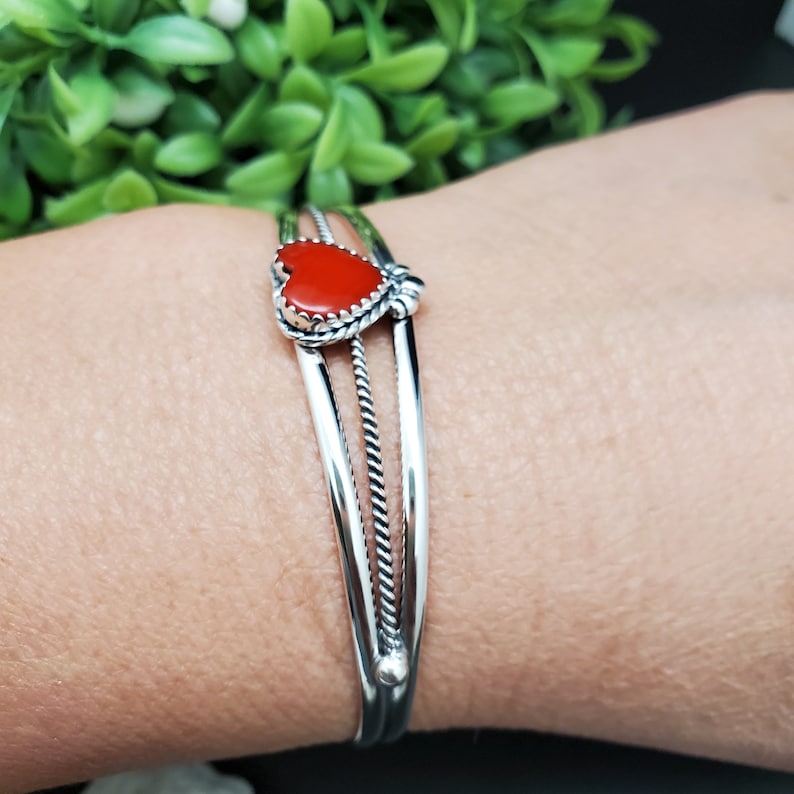 Op de afbeelding: Een zilveren armband met een rode hartvormige steen. De armband heeft drie banden, waarvan &eacute;&eacute;n met een touwontwerp.