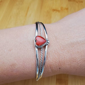 Op de afbeelding: Een zilveren armband met drie banden, waarvan &eacute;&eacute;n gedraaid, en een hartvormige rode koraalsteen in het midden.