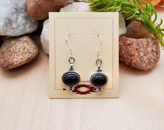 May include: A pair of silver dangle earrings with black oval-shaped stones. The earrings have a simple design with small silver beads around the stones.