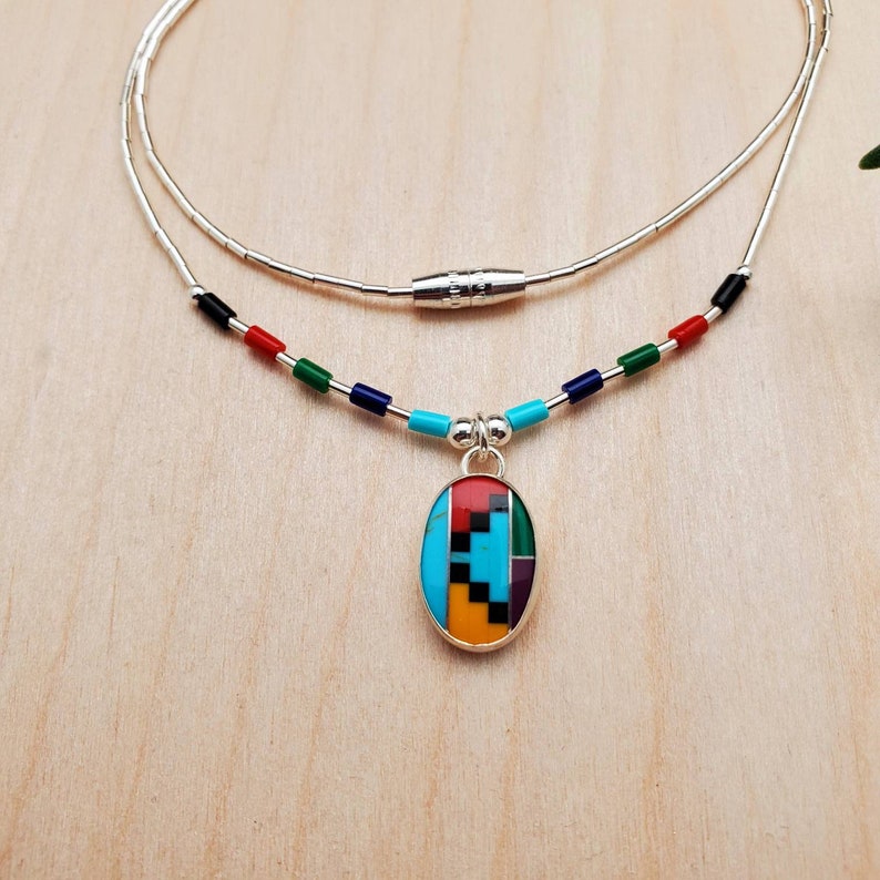 Puede incluir: Un collar de cadena de plata con un colgante ovalado multicolor. El colgante est&aacute; hecho de turquesa, rojo, verde, negro y amarillo. El collar tiene un cierre magn&eacute;tico.