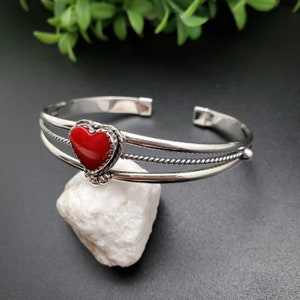 Op de afbeelding: Een zilveren armband met een rode hartvormige edelsteen. De armband heeft een gedraaid draadontwerp en een gladde afwerking.