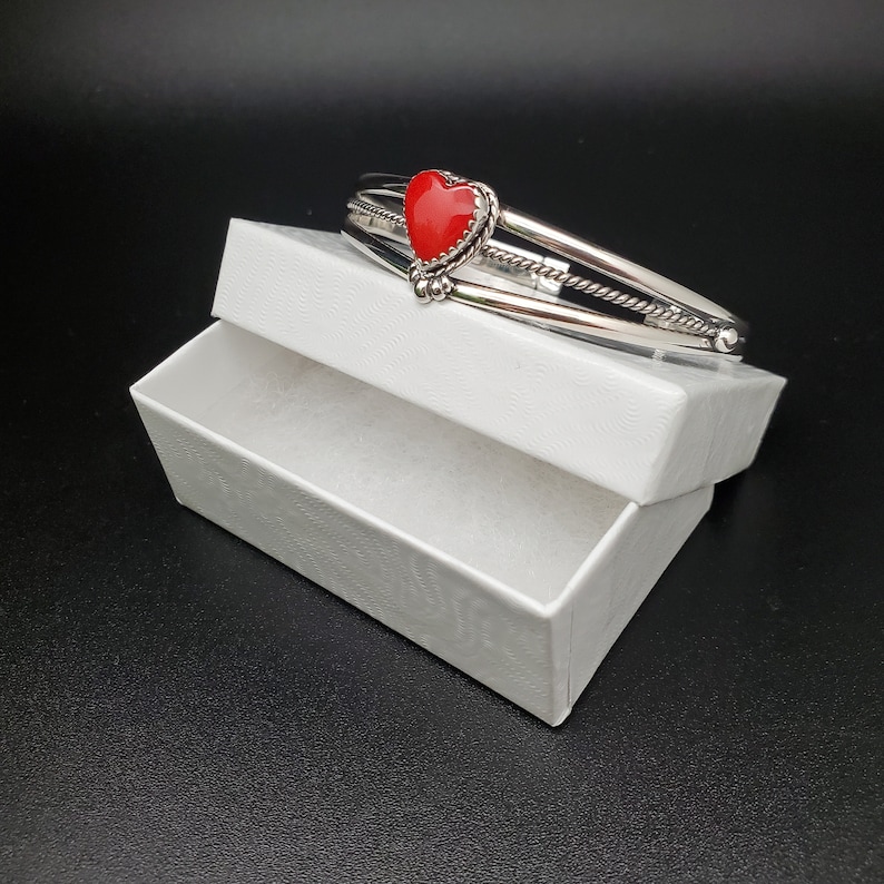 Op de afbeelding: Een zilveren armband met drie banden en een rode hartvormige steen in het midden. De armband zit in een witte doos.