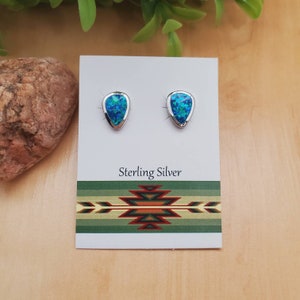 May include: A pair of sterling silver stud earrings with a teardrop-shaped design. The earrings feature a vibrant blue-green opal-like stone set in a silver bezel. The earrings are displayed on a white card with the text "Sterling Silver".