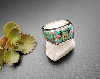 May include: A silver ring with a square shaped stone set in the center. The stone is inlaid with turquoise, coral, and other colorful stones in a geometric pattern.