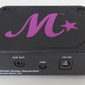May include: Black white noise generator with a purple M and star logo. It has an audio out jack, a power light, and a 12V DC power input. The text on the front reads "White Noise Generator Model WNG-100".