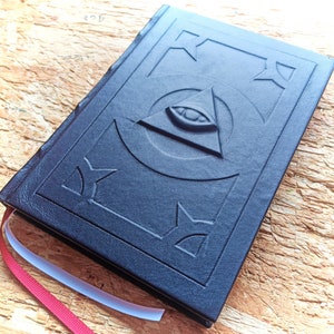 May include: A black leather-bound journal with an embossed all-seeing eye design. The journal has a red ribbon bookmark.