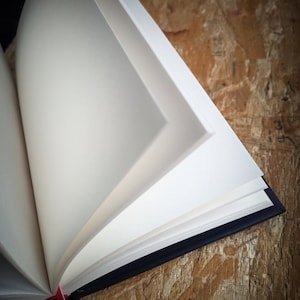 May include: A blank, white notebook with a black cover and a red ribbon bookmark. The notebook is open and lying on a wooden surface.