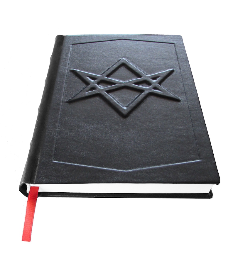 Puede incluir: Un libro encuadernado en cuero negro con un marcap&aacute;ginas de cinta roja. La portada presenta un dise&ntilde;o geom&eacute;trico en relieve.