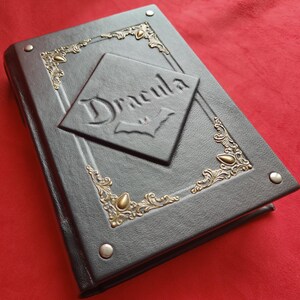 May include: A black leather-bound journal with gold accents and the word "Dracula" embossed on the front cover. The cover features a decorative design with a bat symbol.