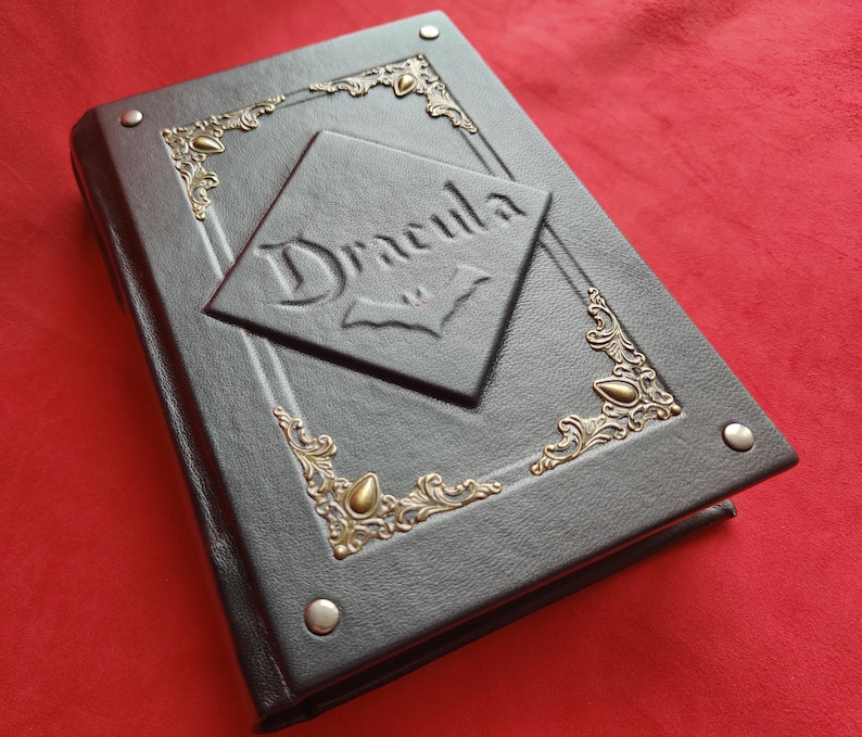 May include: A black leather-bound journal with gold accents and the word "Dracula" embossed on the front cover. The cover features a decorative design with a bat symbol.