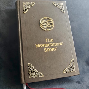 May include: A brown hardcover book titled "The Neverending Story" with gold lettering and decorative corner accents. A gold emblem is centered on the cover. A red and white ribbon bookmark is visible.