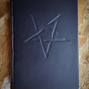 May include: A black leather-bound journal with a pentagram embossed on the front cover. The journal has a red ribbon bookmark.