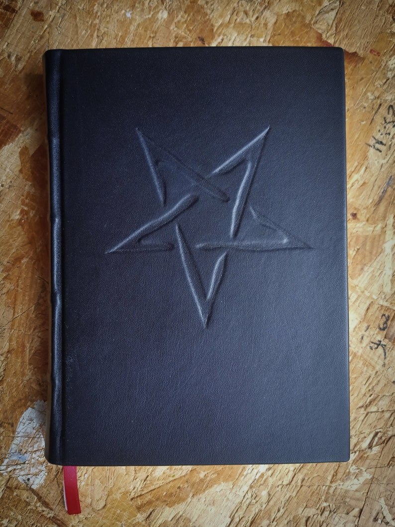 May include: A black leather-bound journal with a pentagram embossed on the front cover. The journal has a red ribbon bookmark.