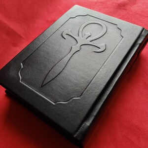 May include: A black leather-bound book with an embossed ankh symbol on the cover.