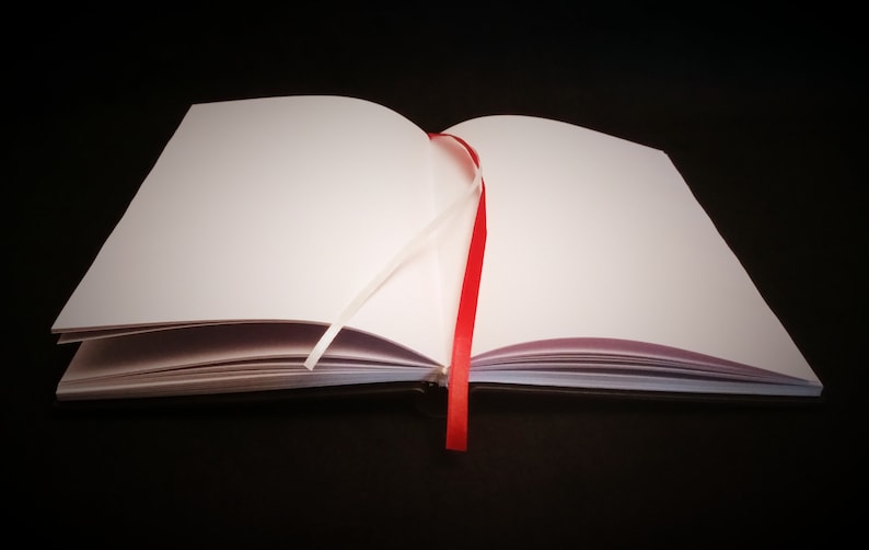 May include: A blank, white notebook with a red ribbon bookmark. The notebook is open and lying flat on a black surface.