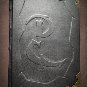 K&ouml;nnte beinhalten: Ein schwarzes, ledergebundenes Tagebuch mit goldfarbenen Metall-Ecken und einem dekorativen, gepr&auml;gten Buchstaben 'E' auf dem Einband.