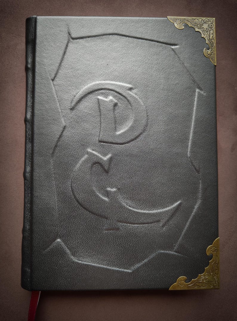 K&ouml;nnte beinhalten: Ein schwarzes, ledergebundenes Tagebuch mit goldfarbenen Metall-Ecken und einem dekorativen, gepr&auml;gten Buchstaben 'E' auf dem Einband.