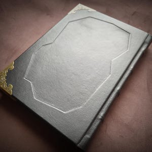 K&ouml;nnte beinhalten: Ein schwarzes, ledergebundenes Tagebuch mit goldfarbenen Metall-Ecken und einem dekorativen, gepr&auml;gten Design auf dem Einband.