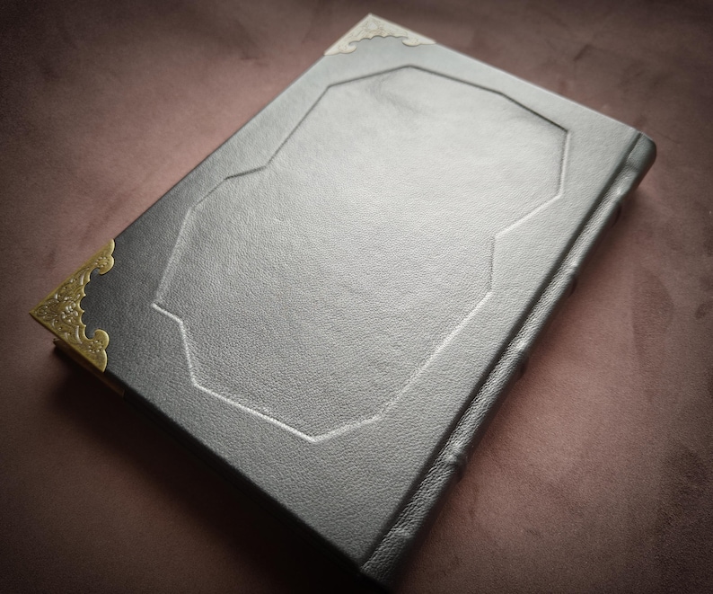 K&ouml;nnte beinhalten: Ein schwarzes, ledergebundenes Tagebuch mit goldfarbenen Metall-Ecken und einem dekorativen, gepr&auml;gten Design auf dem Einband.