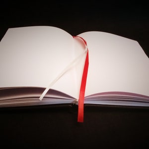 Puede incluir: Un cuaderno blanco en blanco con un marcador de cinta roja. El cuaderno est&aacute; abierto y acostado sobre una superficie negra.