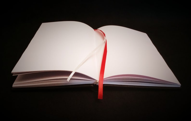 Puede incluir: Un cuaderno blanco en blanco con un marcador de cinta roja. El cuaderno est&aacute; abierto y acostado sobre una superficie negra.