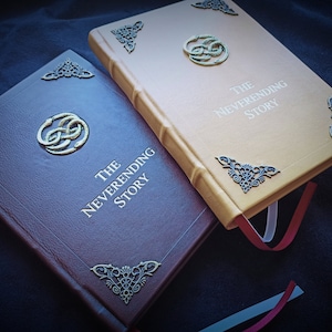 May include: Two hardback books titled "The Neverending Story" are shown. One is brown, the other is tan. Both have decorative metal corner accents and a central emblem. The tan book has a red ribbon bookmark.