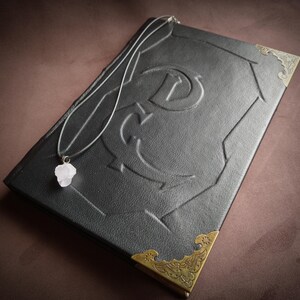 K&ouml;nnte beinhalten: Ein schwarzes, ledergebundenes Tagebuch mit goldenen Verzierungen und einem dekorativen Design auf dem Einband. Eine silberne Kette mit einem rosa Kristallanh&auml;nger liegt auf dem Tagebuch.