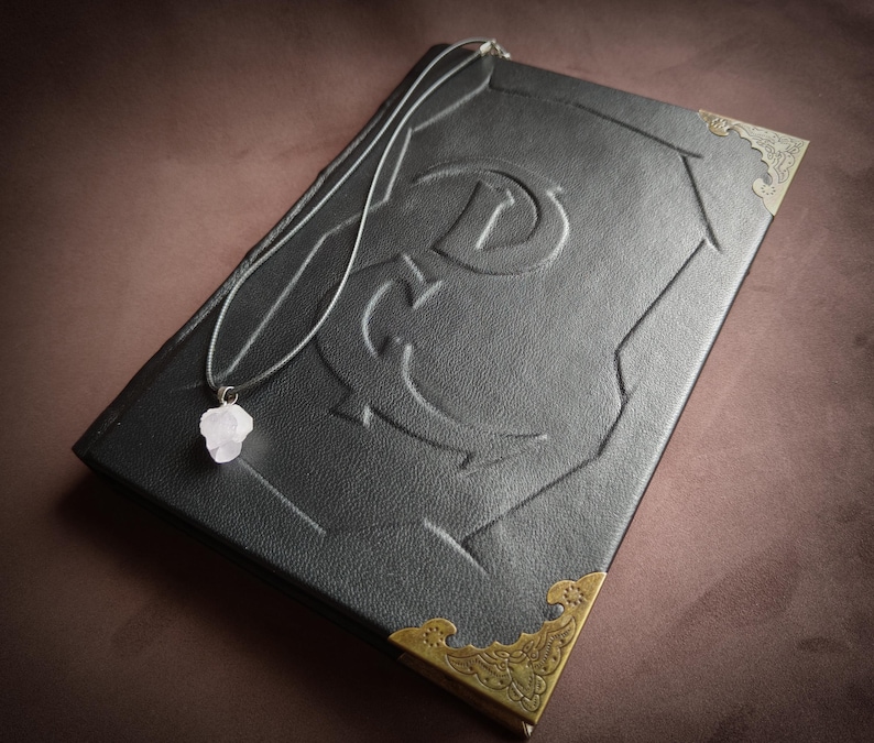 K&ouml;nnte beinhalten: Ein schwarzes, ledergebundenes Tagebuch mit goldenen Verzierungen und einem dekorativen Design auf dem Einband. Eine silberne Kette mit einem rosa Kristallanh&auml;nger liegt auf dem Tagebuch.
