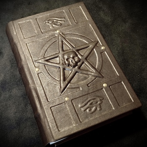 Op de afbeelding: Een donkerbruin, in leer gebonden boek met een in reliëf gemaakt pentagram met een centraal schedelmotief. De kaft bevat twee symbolen van het Oog van Horus en is bevestigd met vier kleine metalen klinknagels op de hoeken van het cirkelontwerp.