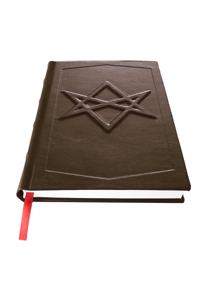Puede incluir: Un libro encuadernado en cuero marr&oacute;n con un marcap&aacute;ginas de cinta roja. La portada presenta un dise&ntilde;o de estrella intrincado en relieve.