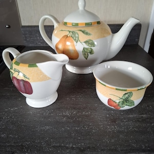 May include: A white ceramic tea set with fruit illustrations. The set includes a teapot with a lid, a creamer, and a small bowl. The teapot features pear illustrations, while the creamer and bowl have cherry and strawberry designs, respectively.