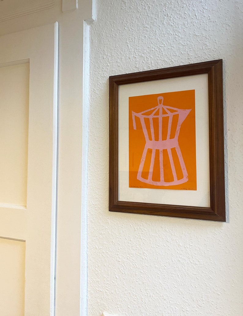 May include: A framed print of a pink coffee pot on an orange background. The print is in a brown wooden frame.
