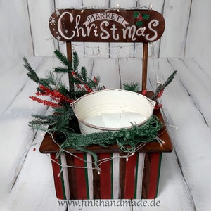 Puede incluir: Un puesto de mercado navide&ntilde;o de madera con un cubo blanco, bayas rojas y ramas de pino verdes. El letrero dice "Mercado de Navidad".