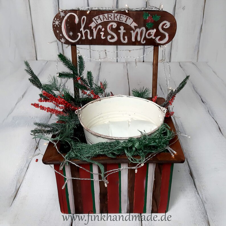 Puede incluir: Un puesto de mercado navide&ntilde;o de madera con un cubo blanco, bayas rojas y ramas de pino verdes. El letrero dice "Mercado de Navidad".