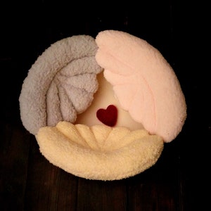 May include: A plush, heart-shaped pillow with three sections in light gray, pale pink, and yellow, surrounding a small red heart. The pillow is made of soft, textured fabric, and is set against a dark background.