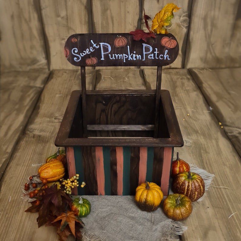 Puede incluir: Una caja de madera con un letrero que dice "Sweet Pumpkin Patch" y est&aacute; decorada con calabazas de oto&ntilde;o. La caja est&aacute; pintada con un patr&oacute;n de rayas en tonos de rosa, verde y marr&oacute;n. Hay varias calabazas peque&ntilde;as alrededor de la caja.