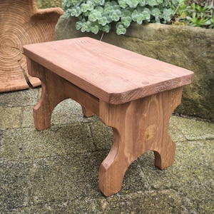 May include: A small wooden stool with a natural brown stain finish. The stool has a rectangular top and four legs with a simple, curved design.