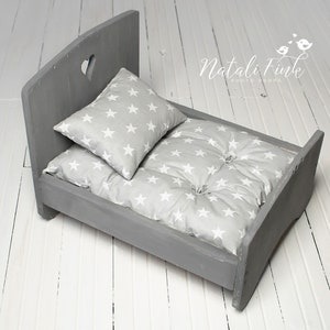 May include: A grey wooden doll's bed with a heart-shaped cutout in the headboard. It features a grey pillow and mattress, both patterned with white stars. The bed is designed for newborn photography, a charming prop.