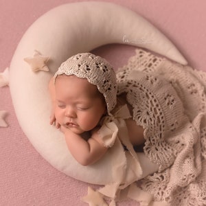 Könnte beinhalten: Ein neugeborenes Baby schläft auf einem weißen, sichelförmigen Kissen, trägt eine Häkelmütze und eine Häkeldecke. Das Baby ist von kleinen weißen Sternen umgeben.