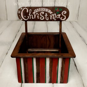 Puede incluir: Un puesto de mercado navide&ntilde;o de madera con una base a rayas rojas, blancas y verdes. El letrero de arriba dice "Mercado de Navidad" con una rama de acebo.