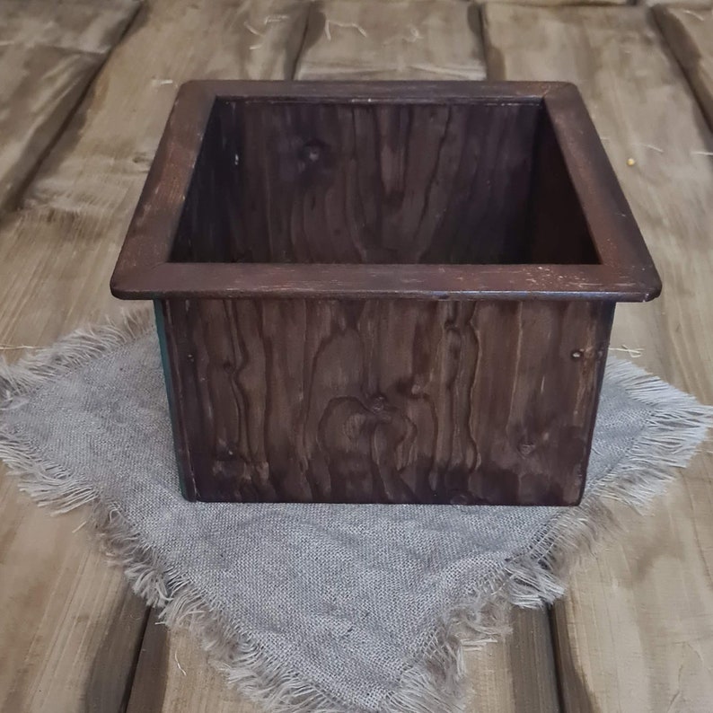 Puede incluir: Una caja de madera marr&oacute;n oscuro con un borde elevado alrededor de la parte superior. La caja est&aacute; sentada sobre un trozo de tela de arpillera.
