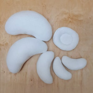 Puede incluir: Una colección de almohadas de felpa blancas, en forma de riñón y circulares, dispuestas sobre una superficie de madera clara. Las almohadas varían en tamaño y forma, con una apariencia suave y texturizada. La disposición sugiere un enfoque en la comodidad y el soporte.