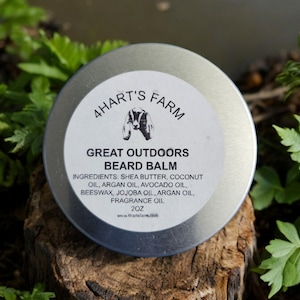 May include: A round, silver tin of "Great Outdoors Beard Balm" from 4Hart's Farm, with a white label. The label includes the brand logo, product name, ingredients, and a 2 oz size. The tin is set on a wooden surface surrounded by green foliage.