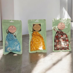 Puede incluir: Tres saquitos de aroma en envases de plástico transparente. Los saquitos están etiquetados como "Clean Linen", "Spiced Pear" y "Christmas Day". Los saquitos están en bolsas de color verde claro con una etiqueta redonda y un cierre de cordón. Los saquitos son azules, amarillos y rojos.