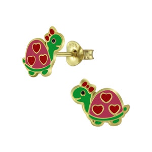 May include: Gold-tone stud earrings featuring a cartoon turtle design. The turtles have green heads, pink shells with red hearts, and red bows. The earrings are set against a white background.