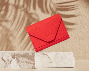 May include: A bright red leather wallet with a fold-over flap and a subtle embossed logo. The wallet is sitting on a white marble surface.