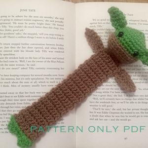 May include: A brown and green crocheted bookmark featuring a character with large eyes and a pointed head. The bookmark is laying on a page of a book with the text "PATTERN ONLY PDF" visible.