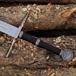 May include: A steel sword featuring a long, sharp blade, a crossguard with twisted metal arms, and a black cord-wrapped handle. The hilt includes two silver-toned metal rings and a circular pommel decorated with a raised, multi-petaled floral emblem.