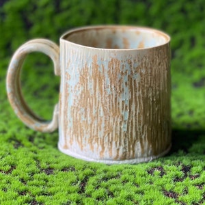 Puede incluir: Una taza de cerámica con un esmalte marrón claro y blanco. La taza tiene una superficie texturizada y un asa.