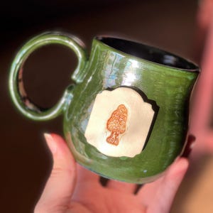 Può includere: Tazza in ceramica fatta a mano con smalto verde lucido. La tazza presenta un emblema rettangolare color crema con un design a fungo in rilievo dettagliato. L'interno è nero e il manico è arrotondato. Un pezzo unico e artistico.